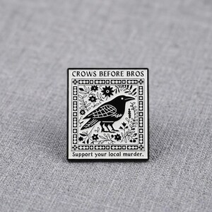 May include: A square enamel pin featuring a black crow centered within a white floral border. The text "CROWS BEFORE BROS" is printed at the top, and "Support your local murder." is printed at the bottom in a black sans-serif font.