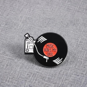 Könnte beinhalten: Eine schwarz-weiße Emaille-Anstecknadel in Form eines Plattenspielers mit einer Vinylplatte. Die Mitte der Platte ist ein leuchtend orangefarbener Kreis mit dem schwarzen Text "MUSIC HEALS MY SOUL". Das Design enthält einen weißen Tonarm.