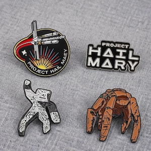 May include: Four enamel pins featuring space-themed designs. One is a circular badge with a spacecraft and the text PROJECT HAIL MARY. Another is a black and white text pin reading PROJECT HAIL MARY. A third is a white and black abstract shape, and the fourth is a brown, multi-legged creature.