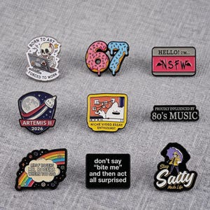 May include: A collection of nine enamel pins featuring various designs including a skeleton at a computer, a doughnut-shaped 67, a name tag reading "HELLO! I'M... NSFW", an Artemis II 2026 space badge, a cat watching a video, and text-based pins.