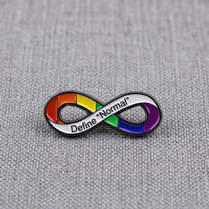 Puede incluir: Un broche de metal con forma de símbolo de infinito, contorno negro y relleno de esmalte con los colores del arcoíris. El texto "Define "Normal"" está impreso en letras negras en el centro del bucle, que presenta segmentos de color rojo, naranja, amarillo, verde, azul y morado.