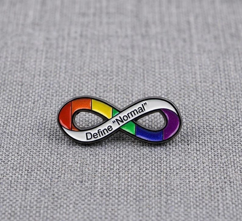 Pu&ograve; includere: Una spilla in metallo a forma di simbolo dell'infinito con contorno nero e smalto color arcobaleno. Il testo "Define "Normal"" &egrave; stampato in lettere nere al centro dell'anello, che presenta segmenti di colore rosso, arancione, giallo, verde, blu e viola.