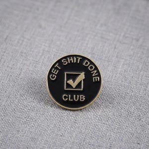 Op de afbeelding: Een ronde metalen speld met een zwarte emaille afwerking en goudkleurige letters en rand. De tekst luidt GET SHIT DONE CLUB, rondom een centraal vierkant vakje met een goudkleurig vinkje, wat zorgt voor een strak en duidelijk ontwerp.