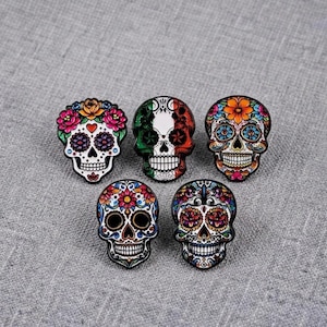 Puede incluir: Cinco pines de esmalte con forma de calavera, cada uno con patrones intrincados y coloridos. Los diseños incluyen motivos florales, formas geométricas y una calavera decorada con los colores verde, blanco y rojo de la bandera mexicana.