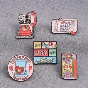 Op de afbeelding: Vijf emaille pins met diverse ontwerpen: een rode telefoon met de tekst I'M HAVING YOUR BABY, een roze ticket met CLUB HEART BREAK, een vierkant bordje IT'S A GOOD DAY TO BE ALIVE, een ronde pin met een hart en een luciferdoosje met TO BE SO LONELY.