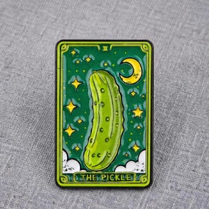 May include: A rectangular enamel pin featuring a green pickle illustration centred on a dark green background with yellow stars and a crescent moon. The bottom of the pin displays the text THE PICKLE in bold yellow capital letters.