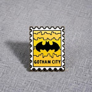 May include: A rectangular enamel pin shaped like a postage stamp with a yellow background, a black bat silhouette in the centre, and a black skyline graphic. The text GOTHAM CITY is printed in black at the bottom of the stamp design.