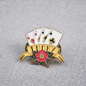 Könnte beinhalten: Eine Metall-Emaille-Anstecknadel mit vier weißen Spielkarten, die die Asse von Herz, Karo, Pik und Kreuz zeigen. Ein gelbes Banner am unteren Rand zeigt das Wort LUCKY in schwarzen Buchstaben, akzentuiert durch eine rote Blume mit grünen Blättern.