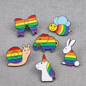 Op de afbeelding: Een verzameling kleurrijke emaille pins met een regenboogstrik, bij, olifant, slak, eenhoorn en konijn. Elke pin heeft een zwarte omtrek en een glanzende afwerking, perfect voor jassen, tassen of hoeden.
