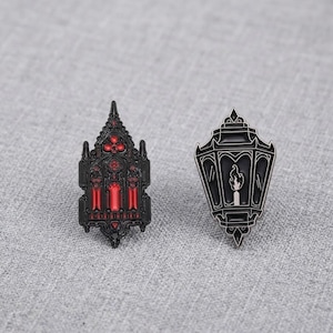 Op de afbeelding: Twee metalen emaille pins. De ene pin is een zwart gotisch kathedraalraam met felrode accenten. De tweede pin is een zwarte lantaarnvorm met een witte kaars en vlam erin, omlijnd met zilverkleurige metalen details.