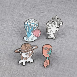 Può includere: Quattro spille smaltate raffiguranti busti stilizzati. Uno è blu e bianco con una bolla rosa. Un altro è bianco con un pezzo di puzzle. Un terzo indossa un cappello a forma di pianeta. L'ultimo è un volto rosa e blu.