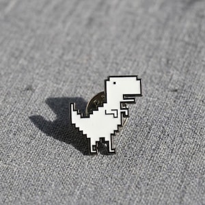 Może przedstawiać: Biała emaliowana przypinka w kształcie spikselowanego dinozaura z czarnymi konturami. Projekt charakteryzuje się blokową estetyką 8-bitową, przedstawiającą widok z boku stojącego stworzenia z małym okiem i wyraźnym ogonem, wykończona metalowym tyłem.