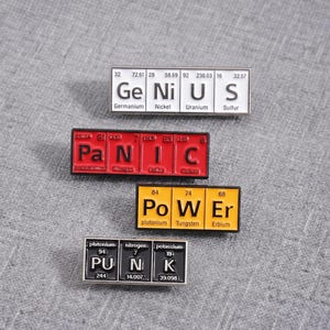 Op de afbeelding: Vier rechthoekige emaille pins met symbolen van elementen uit het periodiek systeem. De bovenste pin is wit met het woord GENIUS, de tweede is rood met PANIC, de derde is geel met POWER en de onderste pin is zwart met het woord PUNK in witte letters.