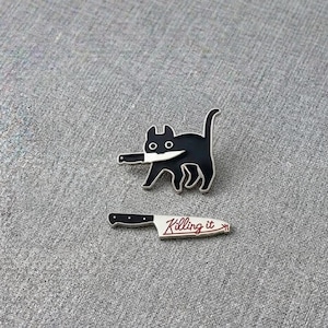 May include: Two enamel pins on a grey surface. One pin is a black cat holding a knife in its mouth. The other pin is a knife with the words "Killing it" in red script.