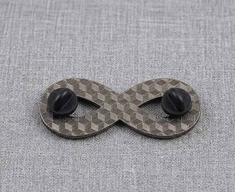 Pu&ograve; includere: Una spilla in metallo a forma di simbolo dell'infinito con un motivo geometrico a cubi testurizzati sulla superficie. Il retro della spilla presenta due chiusure in gomma nera, utilizzate per fissare l'accessorio al tessuto o ad altri materiali.