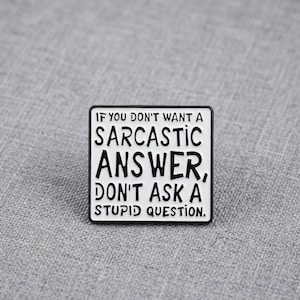 Könnte beinhalten: Eine quadratische Emaille-Anstecknadel mit weißem Hintergrund und schwarzem Rand. Die Anstecknadel zeigt den schwarzen Text: IF YOU DON'T WANT A SARCASTIC ANSWER, DON'T ASK A STUPID QUESTION. Die Schrift ist in einem serifenlosen Stil gehalten.