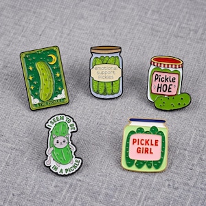 Op de afbeelding: Vijf emaille pins met augurkthema's. Eén is een tarotkaart met het opschrift THE PICKLE, twee zijn potten met het opschrift emotional support pickles en PICKLE GIRL, één pot met het opschrift Pickle HOE, en één is een kat in een augurk met het opschrift I SEEM TO BE IN A PICKLE.
