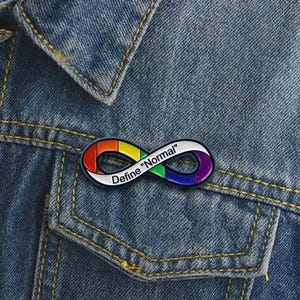 Pu&ograve; includere: Una spilla in smalto a forma di simbolo dell'infinito con un motivo a gradiente arcobaleno sulla superficie. La spilla presenta il testo nero "Define "Normal"" stampato lungo il centro dell'anello, su uno sfondo di tessuto denim blu.