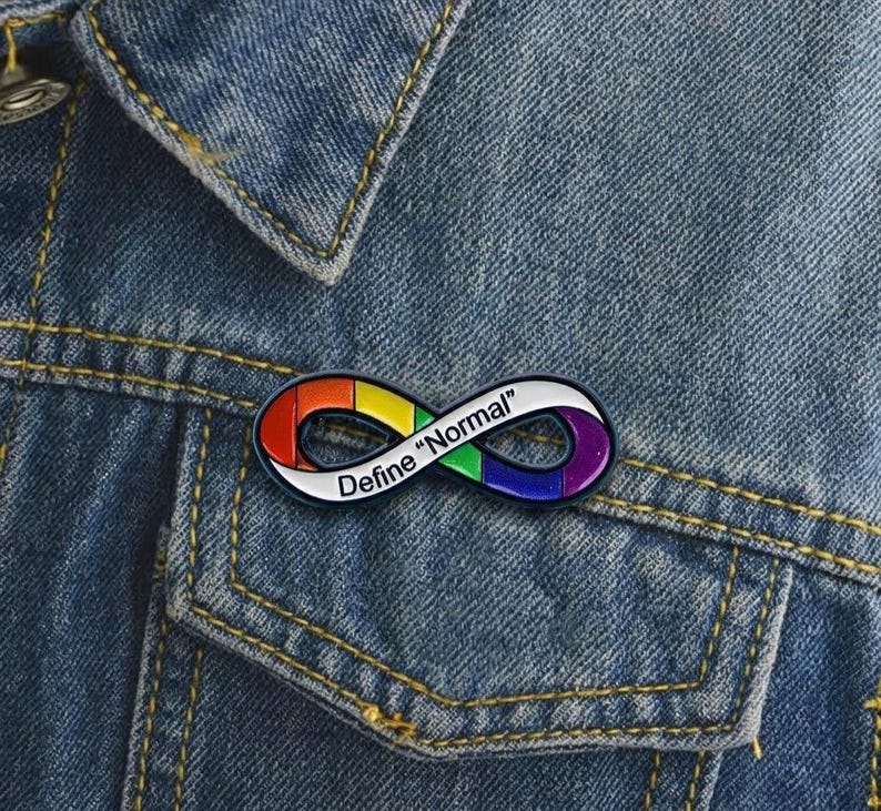 Pu&ograve; includere: Una spilla in smalto a forma di simbolo dell'infinito con un motivo a gradiente arcobaleno sulla superficie. La spilla presenta il testo nero "Define "Normal"" stampato lungo il centro dell'anello, su uno sfondo di tessuto denim blu.