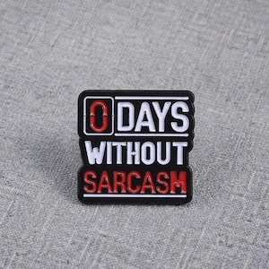 Puede incluir: Un pin esmaltado negro rectangular con el texto blanco "0 DAYS" en la línea superior, "WITHOUT" en la línea central y el texto rojo "SARCASM" en la línea inferior. El número "0" está delineado en rojo y el diseño tiene bordes blancos.