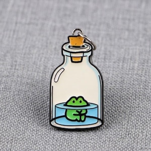 Op de afbeelding: Een metalen emaille speld in de vorm van een doorzichtige glazen fles met een gele kurk. Binnenin de fles zit een kleine groene kikker in een plasje lichtblauw water. De speld is voorzien van een zilverkleurige metalen ring aan de bovenzijde.