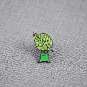 May include: A green enamel pin featuring a cartoon frog holding a large green leaf above its head. The leaf has black text printed on it that reads NEVER FROGET I LOVE YOU, with a small illustration of a snail on the right side of the leaf.