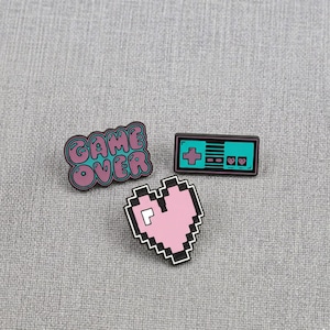 Puede incluir: Tres pines de esmalte con temática de videojuegos. Uno dice "GAME OVER" en letras de burbujas turquesas y rosas. Otro es un corazón rosa pixelado. El tercero es un mando turquesa con borde negro.