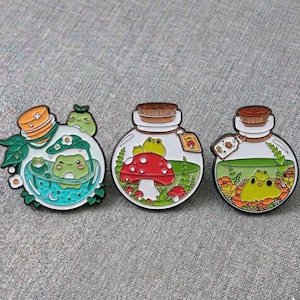 May include: Three enamel pins with quirky designs. One pin depicts frogs in a teal bath, another with a red mushroom, and the last with a frog in a green potion. Each pin is shaped like a jar.