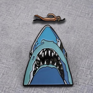 May include: A two-piece enamel pin set featuring a light blue shark head with an open mouth showing sharp white teeth and a separate tan-coloured figure of a person swimming in a horizontal position above the shark head.