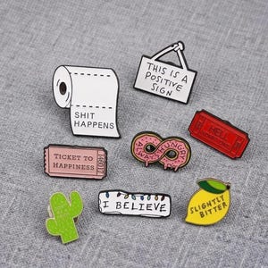 May include: A collection of eight enamel pins featuring various designs: a white toilet paper roll with text, a white sign reading THIS IS A POSITIVE SIGN, a pink donut, a red ticket labeled HELL ADMIT ONE, a pink ticket labeled TICKET TO HAPPINESS, a green cactus, a white banner reading I BELIEVE, and a yellow lemon labeled SLIGHTLY BITTER.