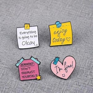 Puede incluir: Cuatro pines de esmalte con forma de notas adhesivas y un corazón. Un cuadrado blanco dice Everything is going to be Okay, un cuadrado amarillo dice enjoy today con un corazón, un cuadrado rosa dice REMINDER: DON'T EXHAUST YOURSELF y un corazón rosa dice I'm fine!