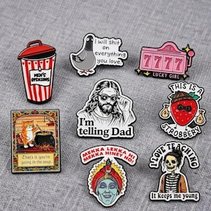 May include: A collection of eight enamel pins featuring various designs including a red and white rubbish bin labelled Men's Opinions, a pigeon, a pink slot machine, a bearded figure, a strawberry, a cat in a soup pot, a genie, and a skeleton.