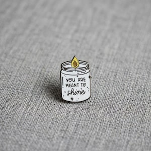 Puede incluir: Un pin de esmalte blanco con forma de vela en tarro con una llama amarilla. La vela presenta el texto negro "YOU ARE MEANT TO SHINE" rodeado de pequeñas estrellas negras. El pin tiene un borde de metal negro y un acabado brillante.