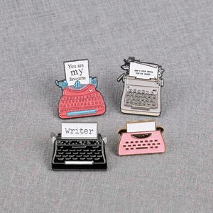 May include: Four enamel pins in the shape of vintage typewriters. The pins are in pink, black, and gray, with white paper inserts. One pin reads "You are my favourite", another says "DON'T STOP UNTIL YOU'RE PROUD", and a third says "Writer".