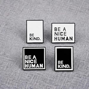 Op de afbeelding: Vier rechthoekige emaille pins met zwart-witte ontwerpen. Twee pins zijn wit met zwarte tekst BE KIND. en BE A NICE HUMAN. Twee pins zijn zwart met witte tekst BE A NICE HUMAN en BE KIND. Alle pins zijn voorzien van dunne randen.