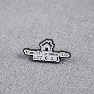 Peut inclure: Une broche en émail noir et blanc en forme d'icône de maison au-dessus du texte "There is no place like 127.0.0.1". Le design présente un fond blanc avec des lettres noires et un contour noir, créant un graphique contrasté pour cet accessoire.