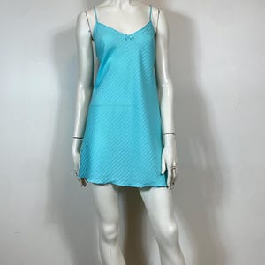 K&ouml;nnte beinhalten: Ein hellblaues, &auml;rmelloses Schlupfkleid mit einem d&uuml;nnen wei&szlig;en Streifenmuster. Das Kleid hat einen V-Ausschnitt und eine Schleife am Ausschnitt.