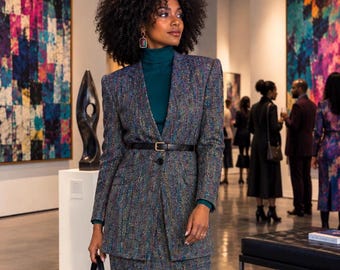 May include: A dark gray tweed blazer and matching skirt set featuring multicolored flecks, worn over a teal turtleneck sweater. The blazer is cinched at the waist with a thin black leather belt with a gold buckle, and a black handbag is held.