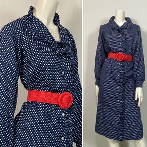 Pu&ograve; includere: Un abito a pois blu navy e bianco con colletto a volant e maniche lunghe. L'abito ha una cintura rossa con una grande fibbia rotonda.