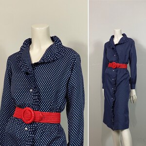 Pu&ograve; includere: Un abito blu navy a pois con colletto a volant e maniche lunghe. L'abito ha una cintura in tessuto rosso con una fibbia grande.