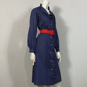 Pu&ograve; includere: Un abito blu navy con pois bianchi e un colletto a balze. L'abito ha una chiusura con bottoni sul davanti e maniche lunghe. Una cintura rossa con una grande fibbia viene indossata in vita.