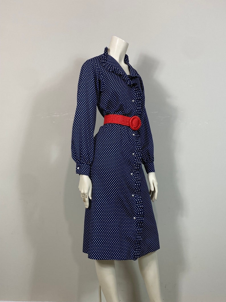 Pu&ograve; includere: Un abito blu navy con pois bianchi e un colletto a balze. L'abito ha una chiusura con bottoni sul davanti e maniche lunghe. Una cintura rossa con una grande fibbia viene indossata in vita.