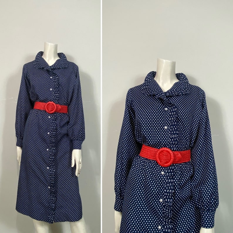 Pu&ograve; includere: Un abito a pois blu navy con colletto a volant e maniche lunghe. L'abito ha una chiusura con bottoni sul davanti e una cintura rossa.