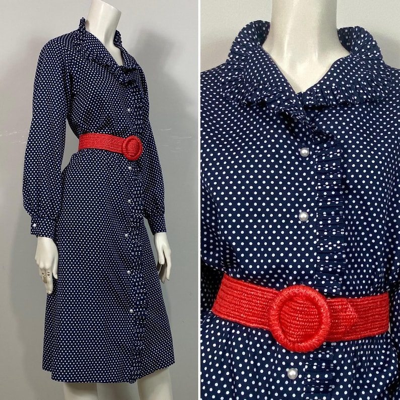 Pu&ograve; includere: Un abito blu navy con pois bianchi e un colletto a balze. L'abito ha una chiusura con bottoni sul davanti e una cintura rossa con una grande fibbia circolare.