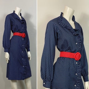 Pu&ograve; includere: Un abito blu navy con pois bianchi e un colletto a balze. L'abito ha maniche lunghe e una chiusura con bottoni sul davanti. &Egrave; stretto in vita da una cintura rossa.