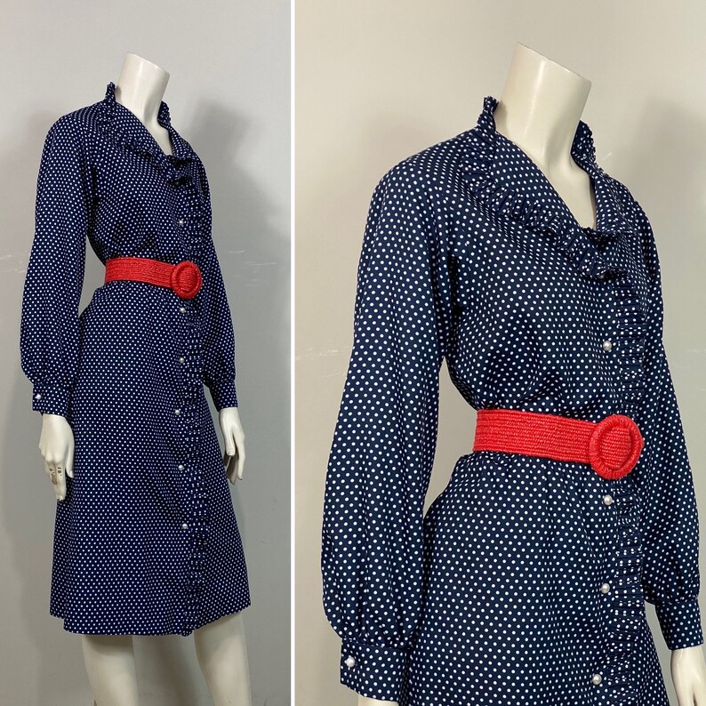 Pu&ograve; includere: Un abito blu navy con pois bianchi e un colletto a balze. L'abito ha maniche lunghe e una chiusura con bottoni sul davanti. &Egrave; stretto in vita da una cintura rossa.