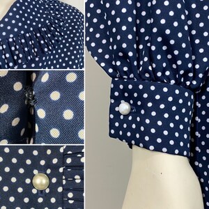 Pu&ograve; includere: Un abito blu navy con pois bianchi e bottoni di perla. L'abito ha una scollatura arricciata e maniche lunghe con polsini arricciati.