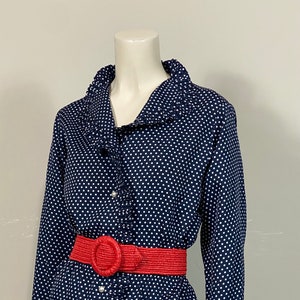Pu&ograve; includere: Una camicia a maniche lunghe blu navy con pois bianchi e un colletto arricciato. Una cintura intrecciata rossa con una grande fibbia circolare &egrave; indossata in vita.