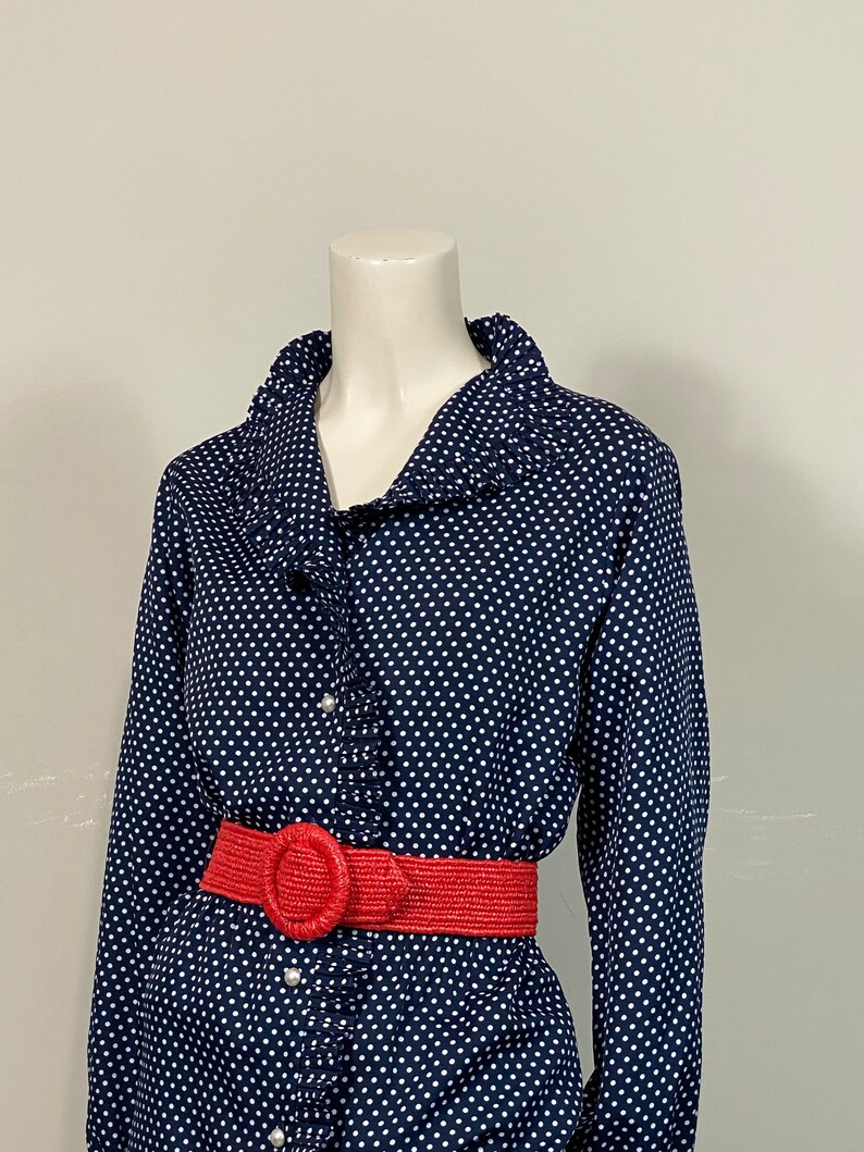 Pu&ograve; includere: Una camicia a maniche lunghe blu navy con pois bianchi e un colletto arricciato. Una cintura intrecciata rossa con una grande fibbia circolare &egrave; indossata in vita.