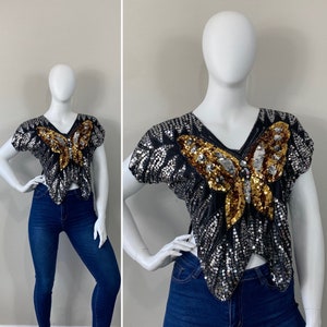 Puede incluir: Una blusa negra y plateada con lentejuelas y un diseño de mariposa dorada. La blusa tiene cuello en V y mangas cortas.
